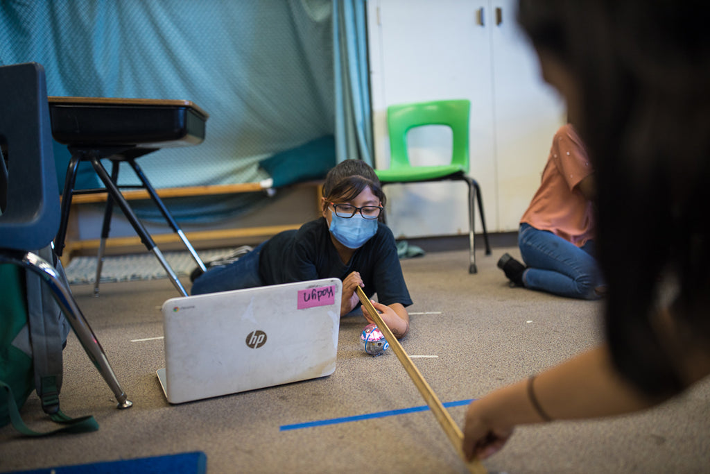 A girl wearing a mask measures distance with a yard stick while coding her Sphero robot with a laptop.