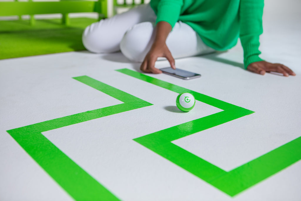 A green Sphero Mini programmable robot navigates a tape maze on the floor.