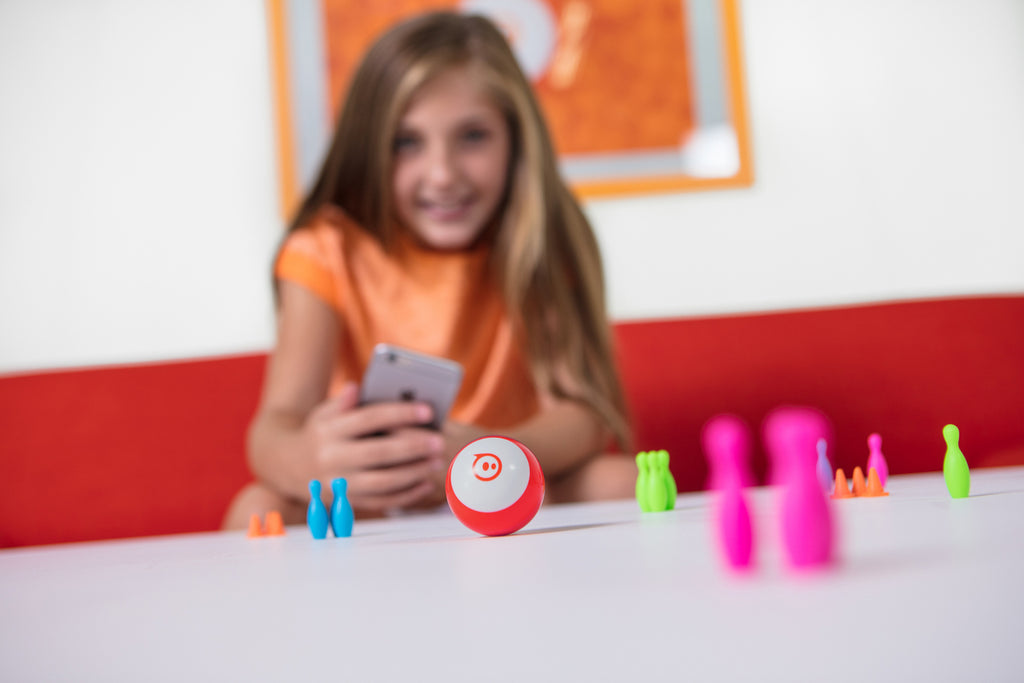 A girl programs her Sphero Mini through bowling pins and cones on a table inside.