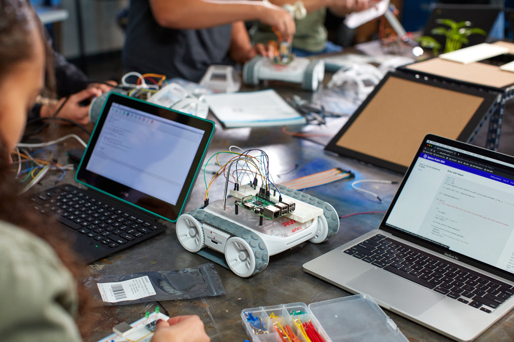 A table full of Sphero RVR+ robots and laptops containing text code.