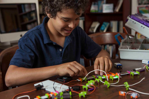 A boy builds a littleBits invention at a table in a classroom.