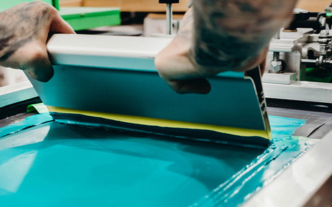 person pulling a squeegee across a screen filled with light blue ink