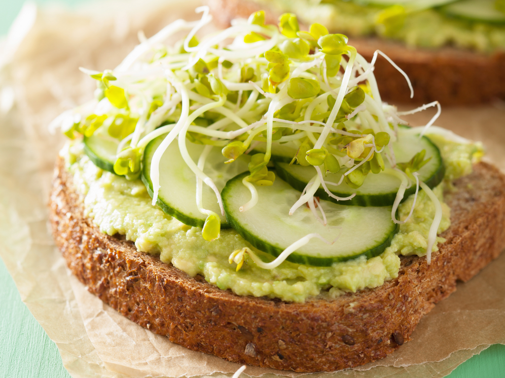 Slice of seed bread with guacamole, cucumbers, and seed sprouts on a table.