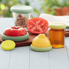 Set of four foodhuggers that preserve a tomato, a lemon and also a replacement lid for jars, on a wooden table.