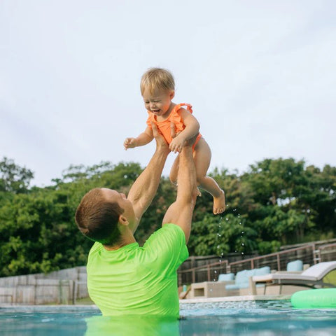 Girl in a SwimZip neon orange one-piece swimsuit plays with her dad in the swimming pool—What is high-visibility swimwear?