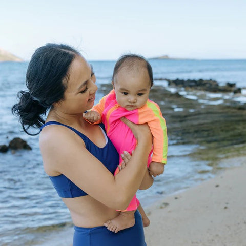 Kid in a SwimZip neon pink and orange sunsuit with their mom at the beach—What is high-visibility swimwear?