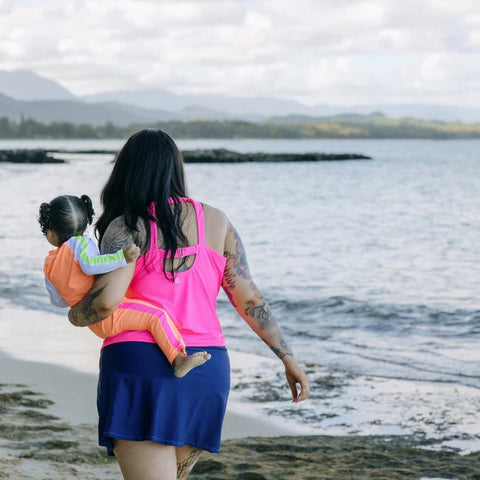 Woman in a SwimZip neon pink tankini top walks along the beach carrying her child—What is high-visibility swimwear?