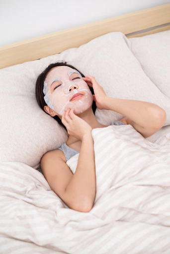 girl lying down on the bed with white sheet mask and touching her face