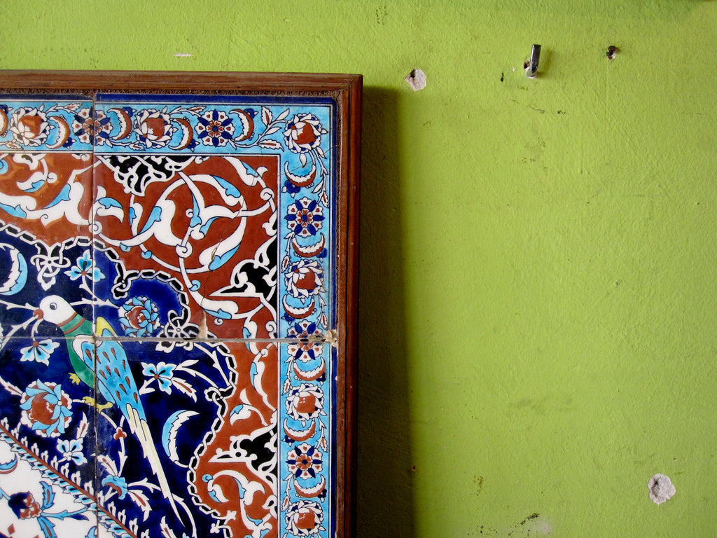 Photo of a traditional Turkish tile art in blues, white and brown with detail of a bird and flowers hung on a pistachio coloured wall.