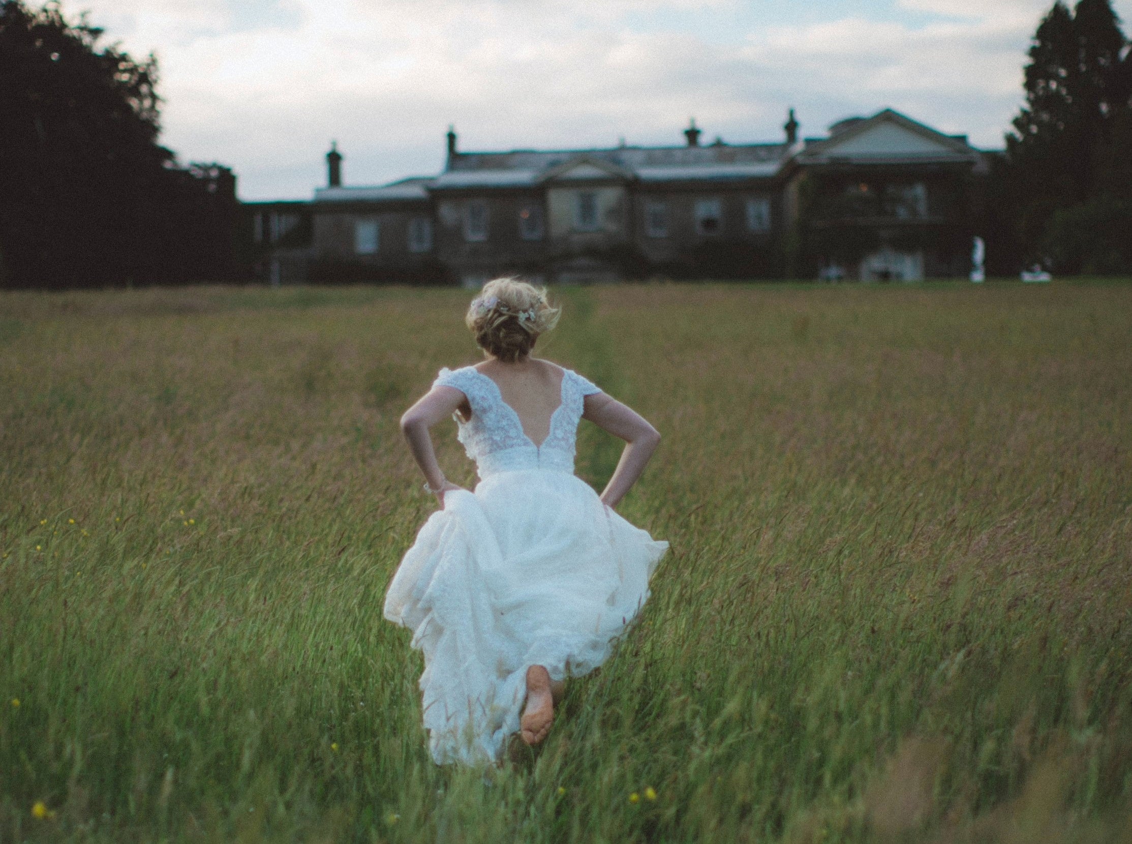 Woman in wedding dress runs through field towards large country house