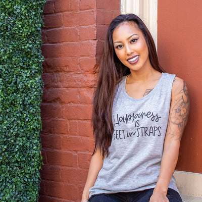 Woman wearing a grey muscle tank top that says "happiness is feet in straps"