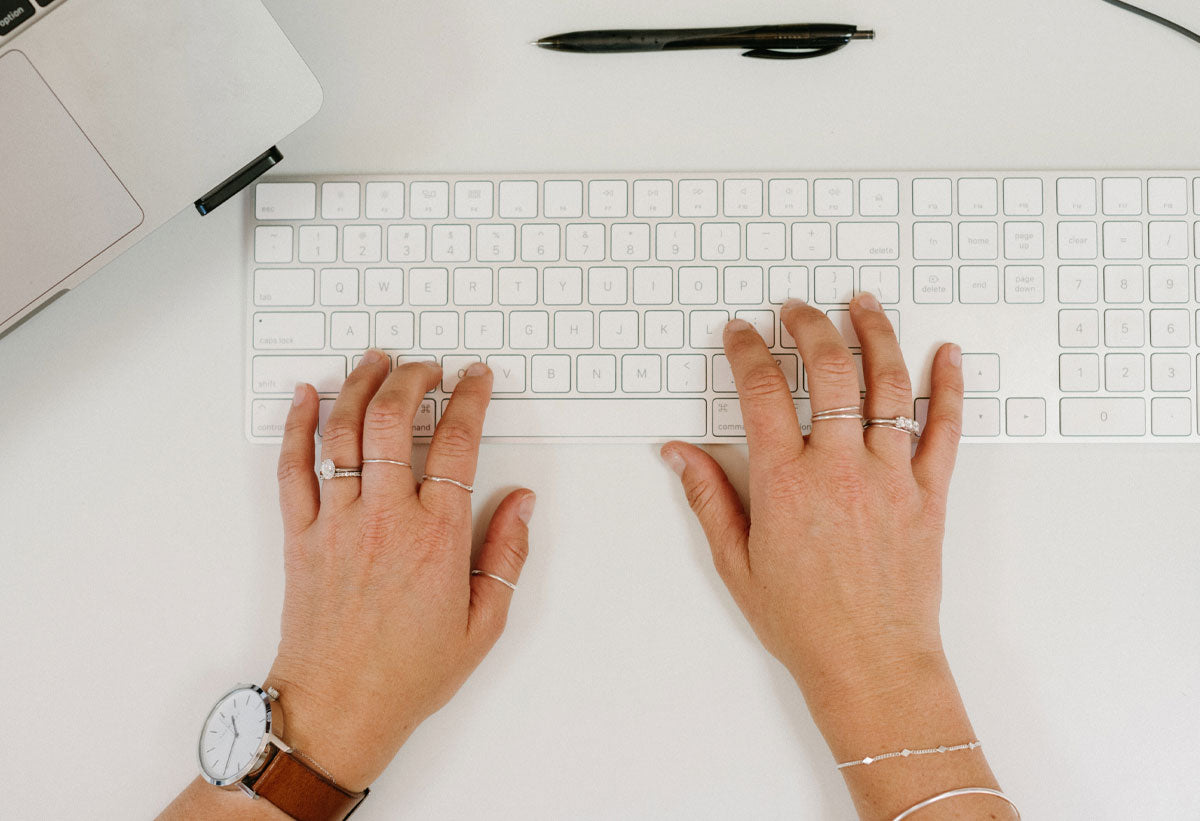 Kristen Sutherland's hands typing on a grey Mac keyboard wearing White Gold rings