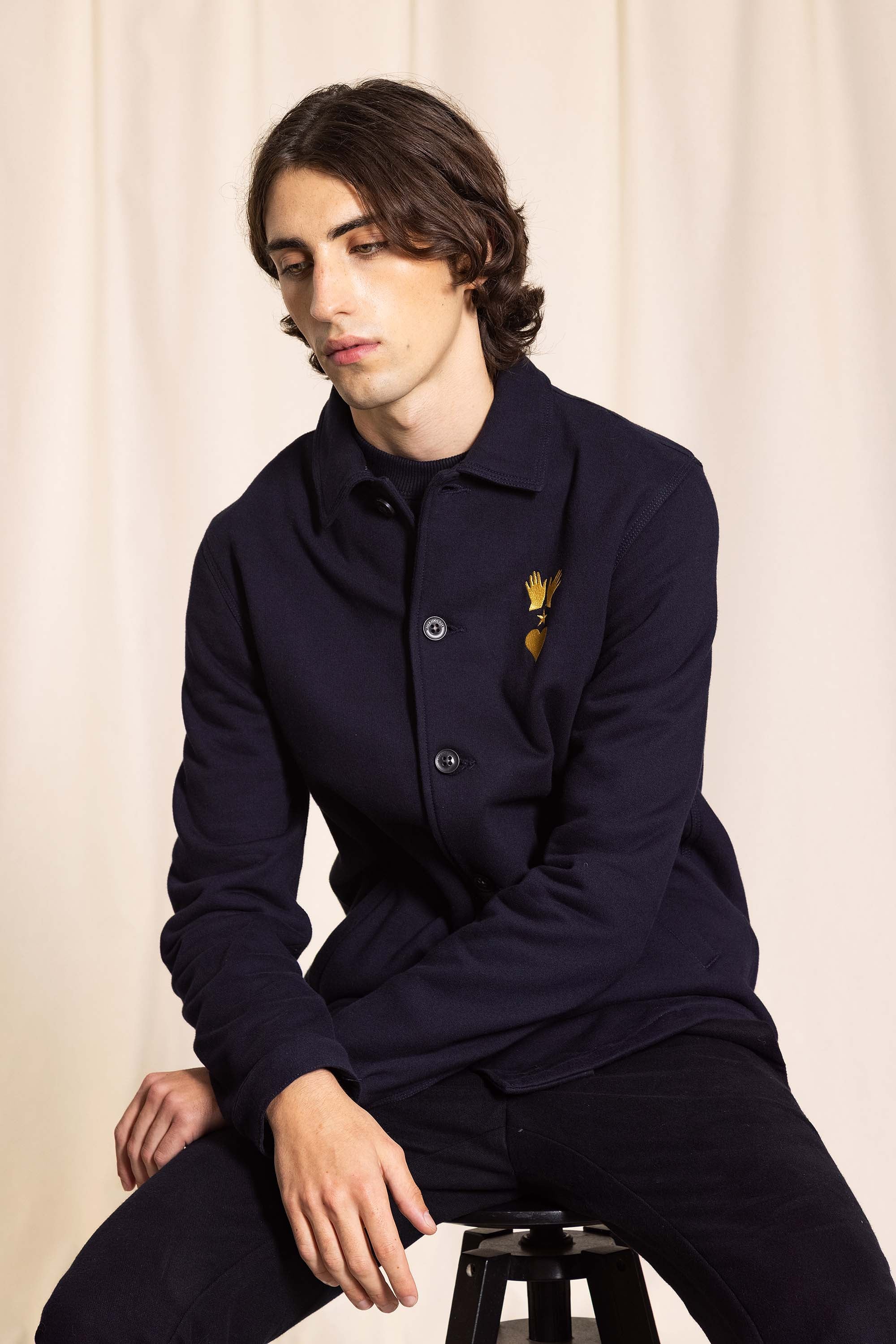 seated man wearing a navy estevez jacket buttoned and embroidered on the chest, worn with shirts t-shirts or under-sweaters