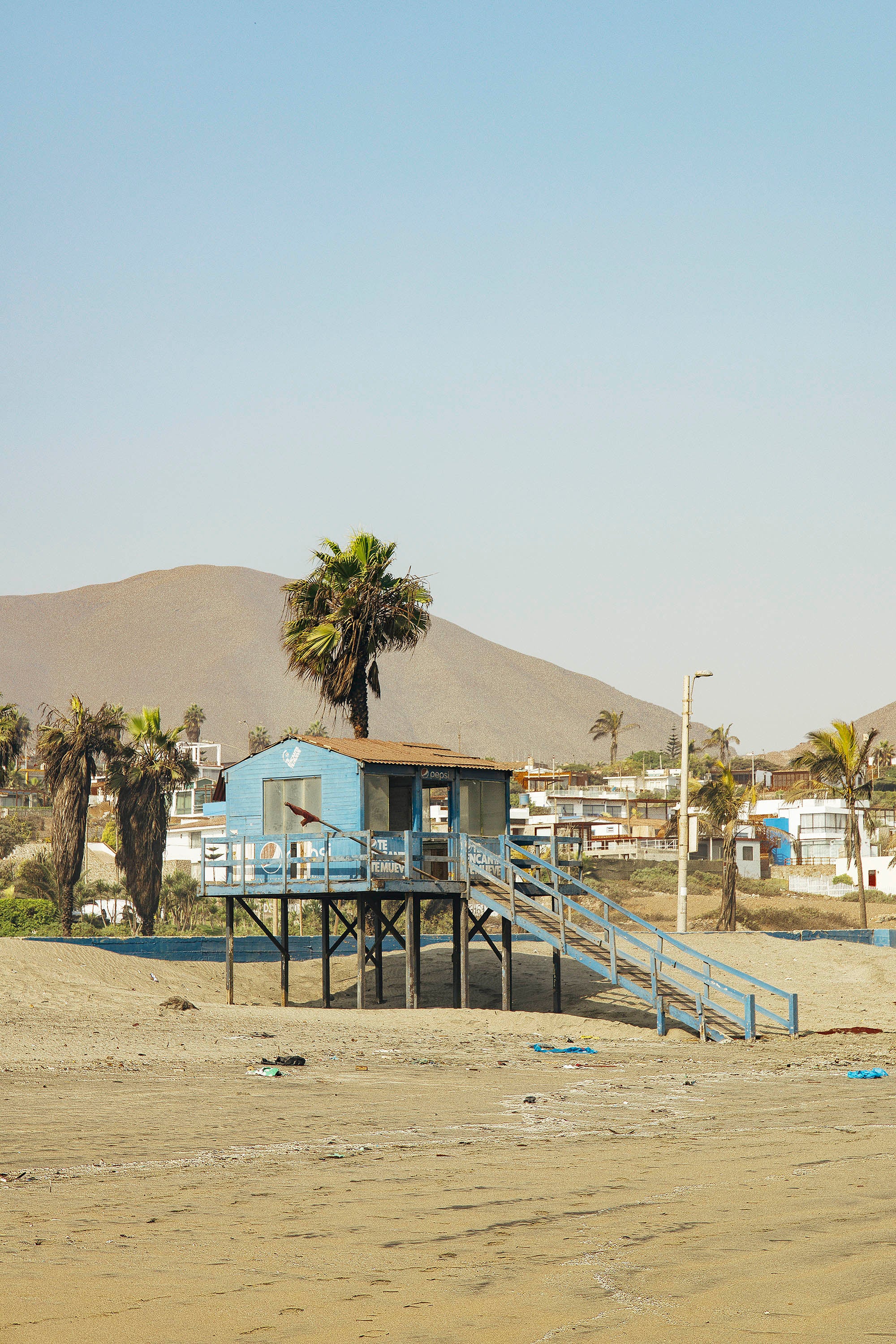 Cabanon sur la plage photographié avec palmiers et habitations visibles en arrière plan ainsi que les montagnes au pérou