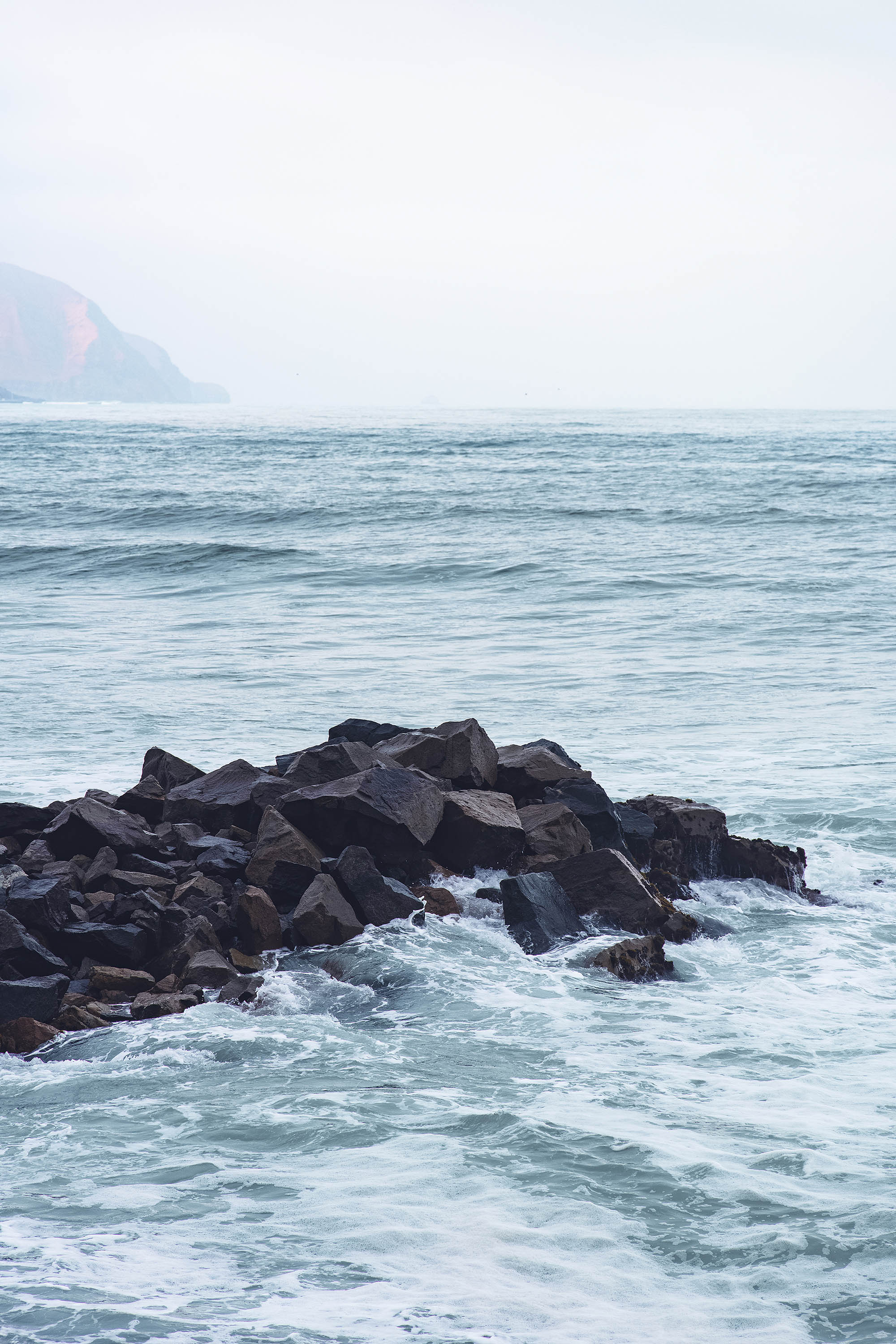 Rocks by the ocean photographed under a misty sky with a mountain visible in the background Lima Peru Landscape Slums city light architecture desert wave