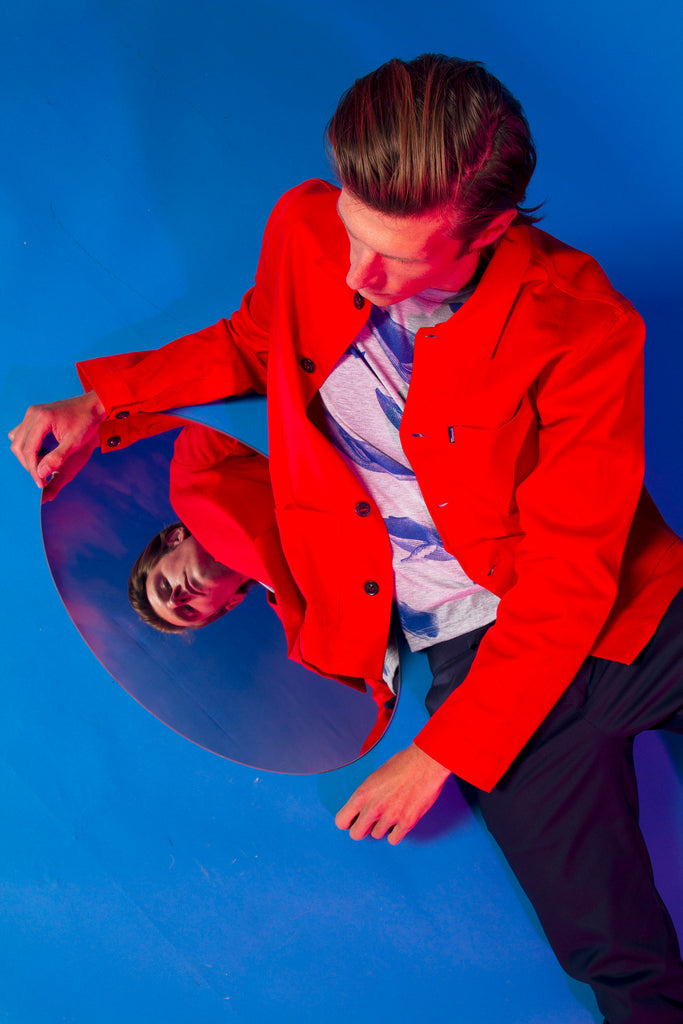 Blond model seated on the floor in a red jacket like buttoned work blue in cotton, straight cut with three pockets on the front utilitarian garment workwear inspiration for an ideal Parisian dressing room