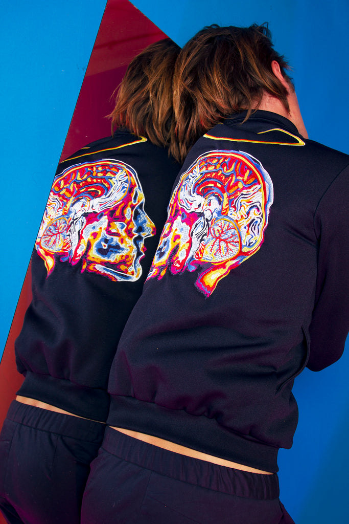 Man lying on a mirror wears a midnight blue sports jacket in cotton and polyester with skull embroidery on the back Lima fair trade spiritual and scientific