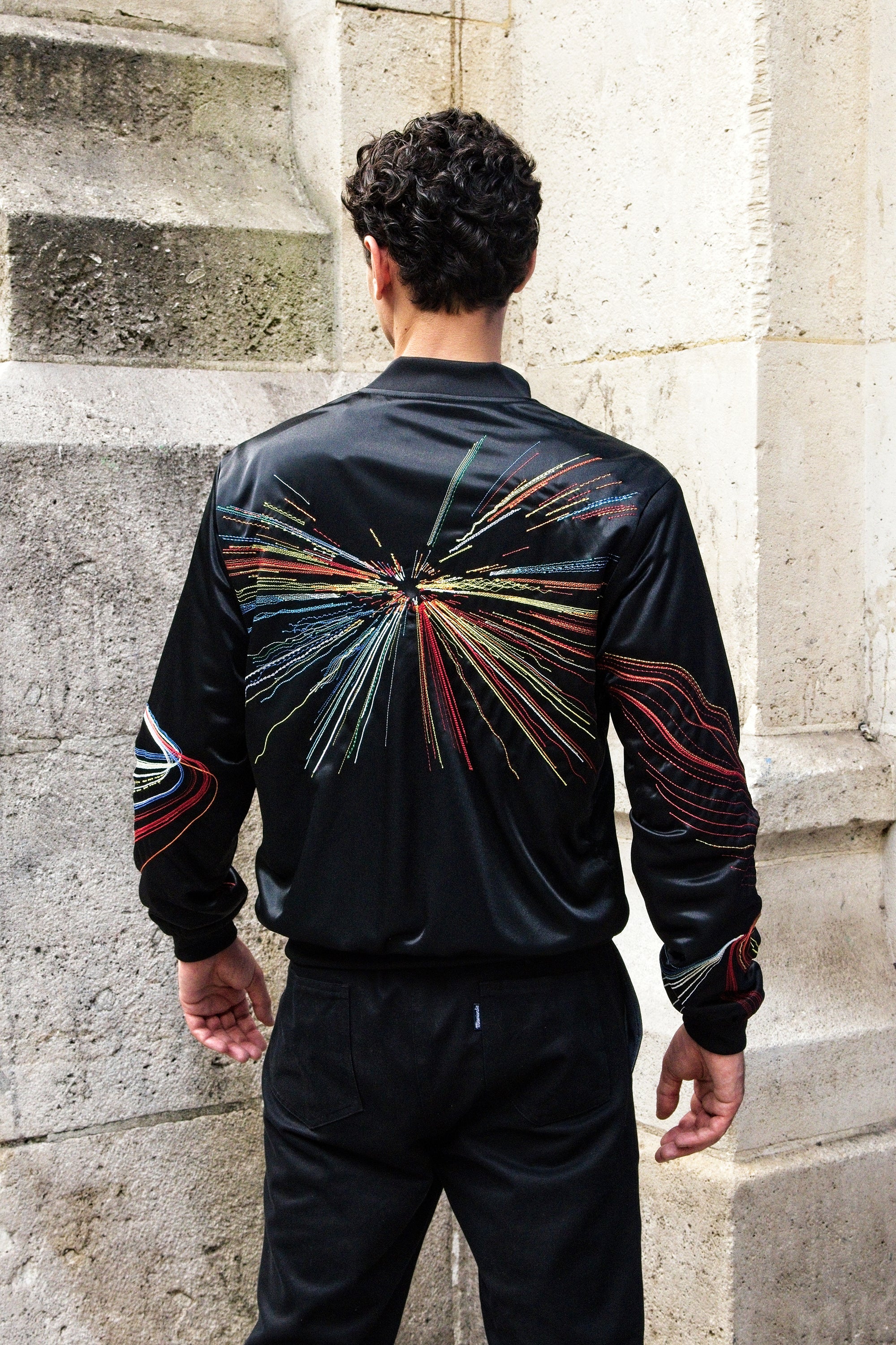Man from behind wearing a black satin souvenir jacket. The embroidery represents the light of headlights at night, it is found on the back as well as on the sleeves
