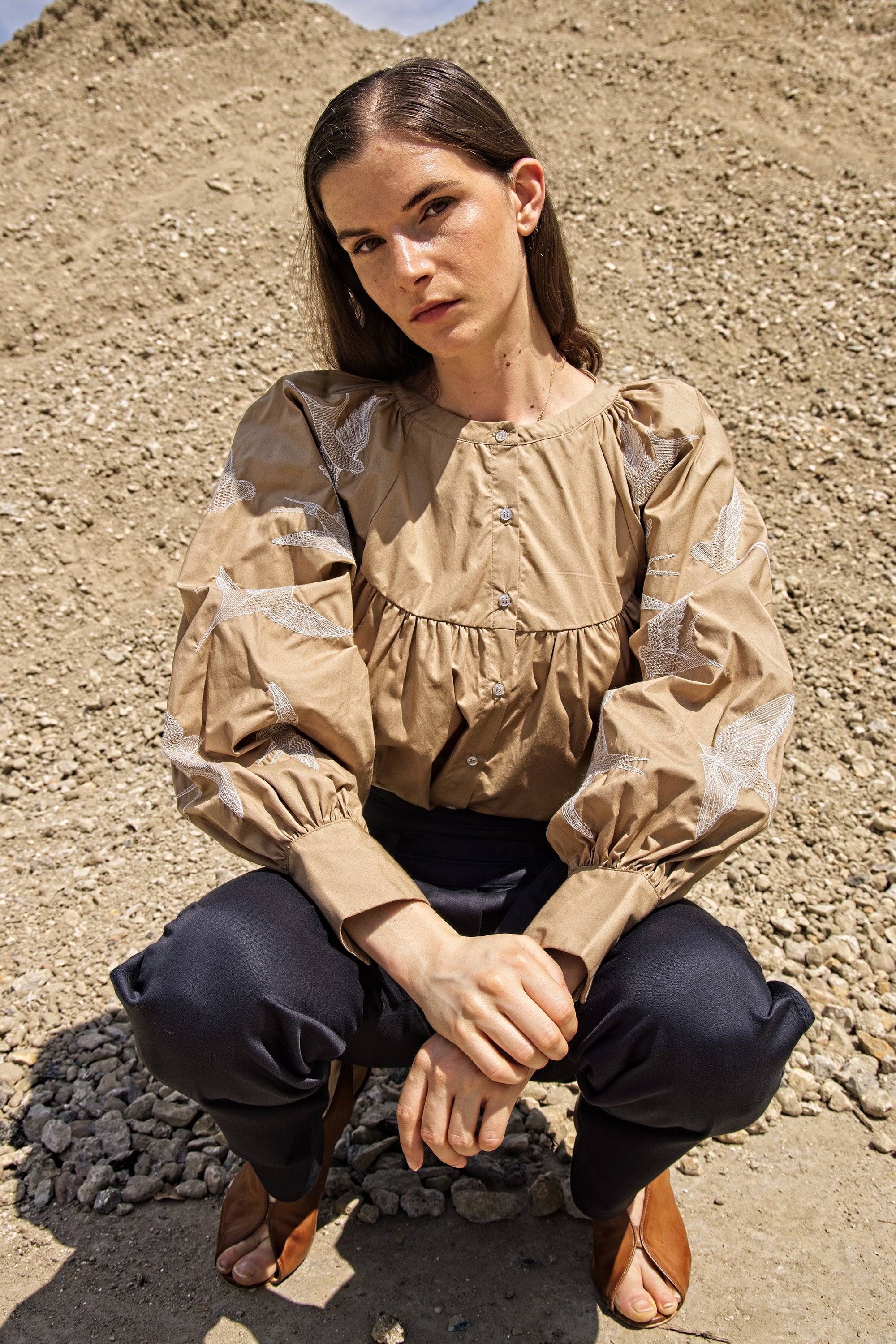 Femme portant une chemise beige avec des broderies sur les manches. La chemise est en coton , courte et ample, elle est boutonnée avec un col rond . Les manches raglant sont brodés en blanc de gros oiseaux en vol.
