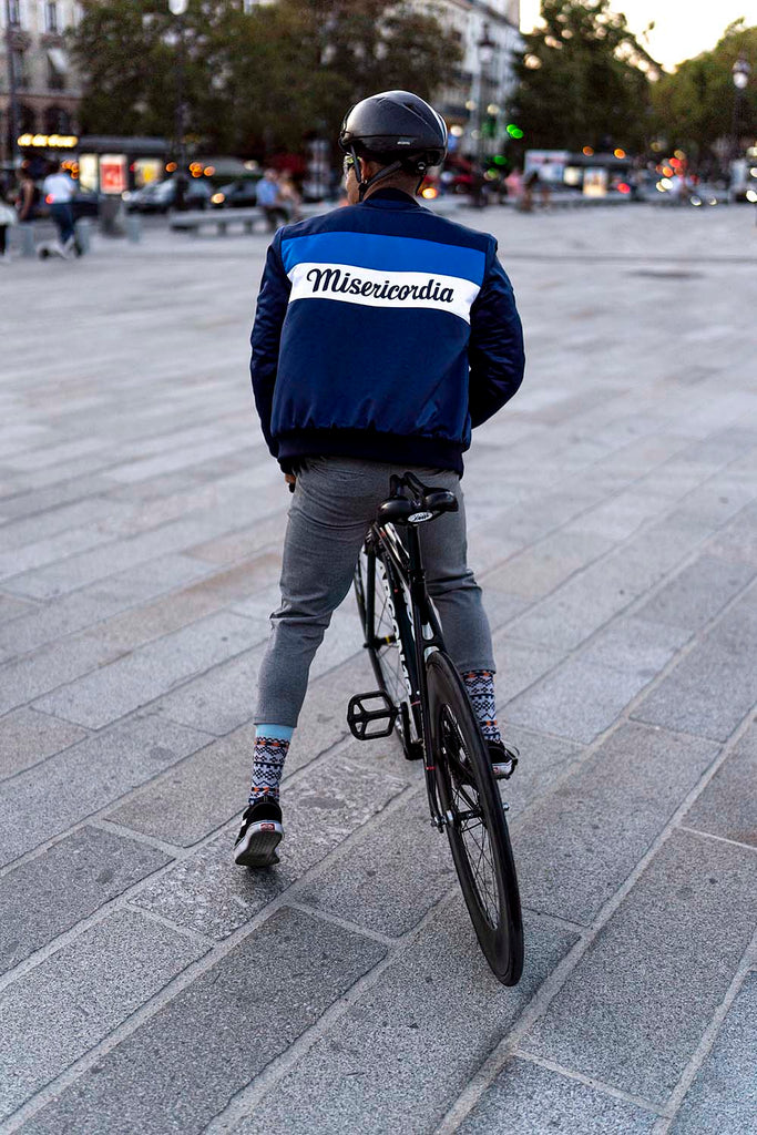 Man from the back, stationary with his bike, sporty chic wearing a satin teddy jacket bearing the image of the responsible brand Misericordia, delivery by bike delivery man Paris and small belt