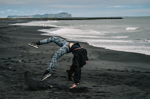 Imagen de una mujer saltando hacia atrás en una playa de arena negra con pantalones de yoga grises.