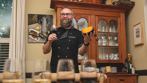 Andrew Wheeler from TOMME Cheese Shop at a wine and cheese event. He is holding a glass of red wine and an orange wedge of Mimolette cheese.