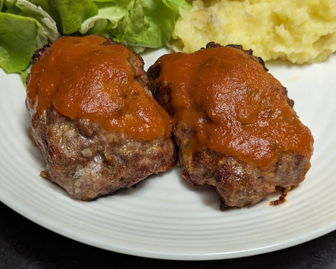 Two mini meatloaves glazed with ketchup on a plate with mashed potatoes and salad greens.