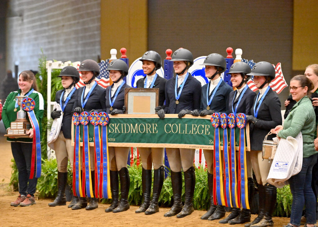 8 female equestrian competitors holding a "Skidmore College" banner with winning ribbons