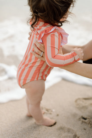 A baby is held above the sand wearing a UPF 50 sun protective swimsuit.