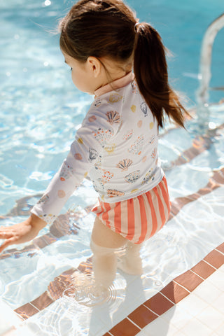 A little girl stands on the steps of a pool in Pehr’s Seashell Rash Guard and Striped Bottom.