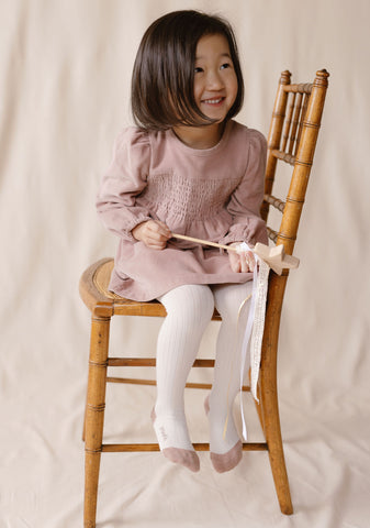 An image of a young girl wearing a pink corduroy dress and tights and sitting on a wooden chair.