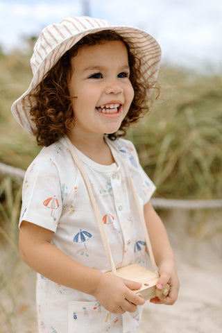 A girl, wearing a By The Sea romper and bucket hat, stands on the beach with a toy camera.