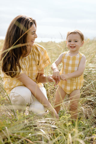 A mom and her son, wearing matching Checkmate clothing, smile at each other while standing in tall grass.