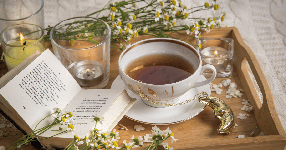 Chamomile tea and flowers with candles, petals and book on a bed tray.