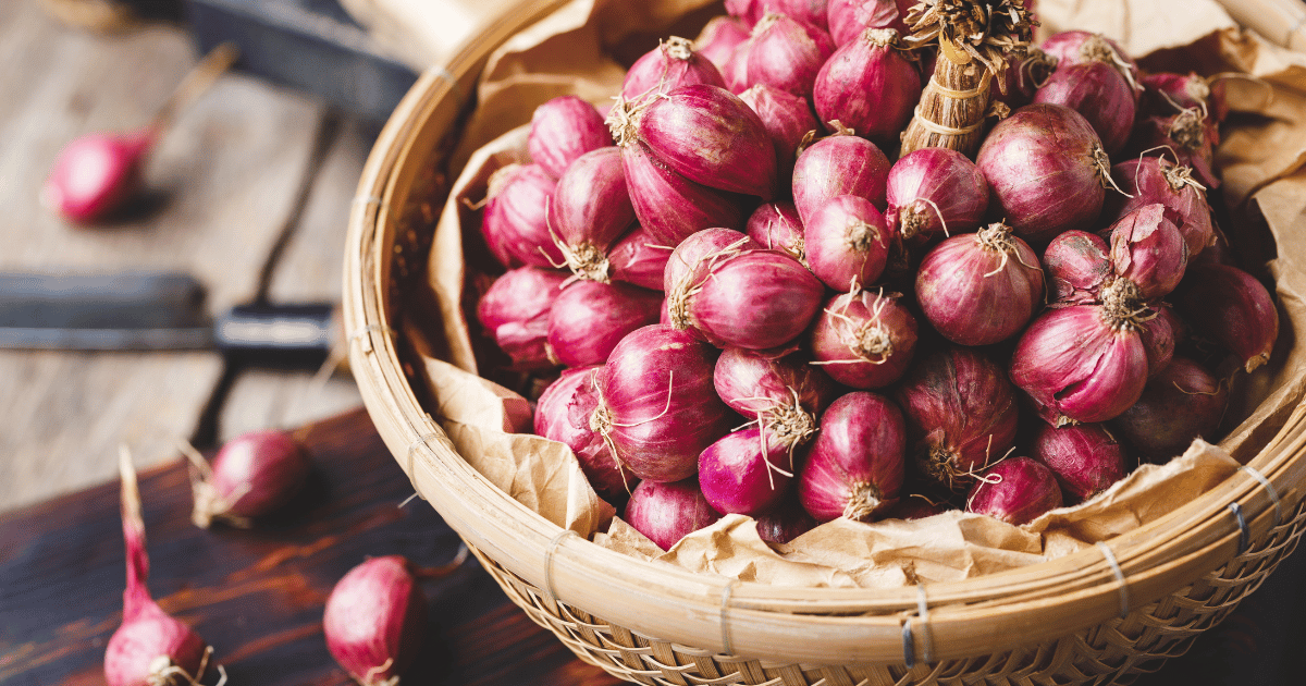 A basket of shallots with a knife in the background on a wood table.