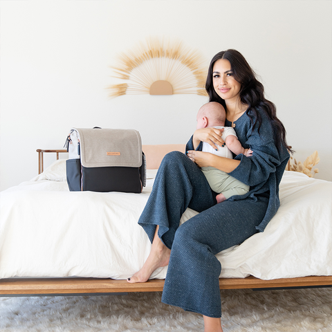 Mother sitting on bed, holding baby, next to Petunia Pickle Bottom Diaper Bag