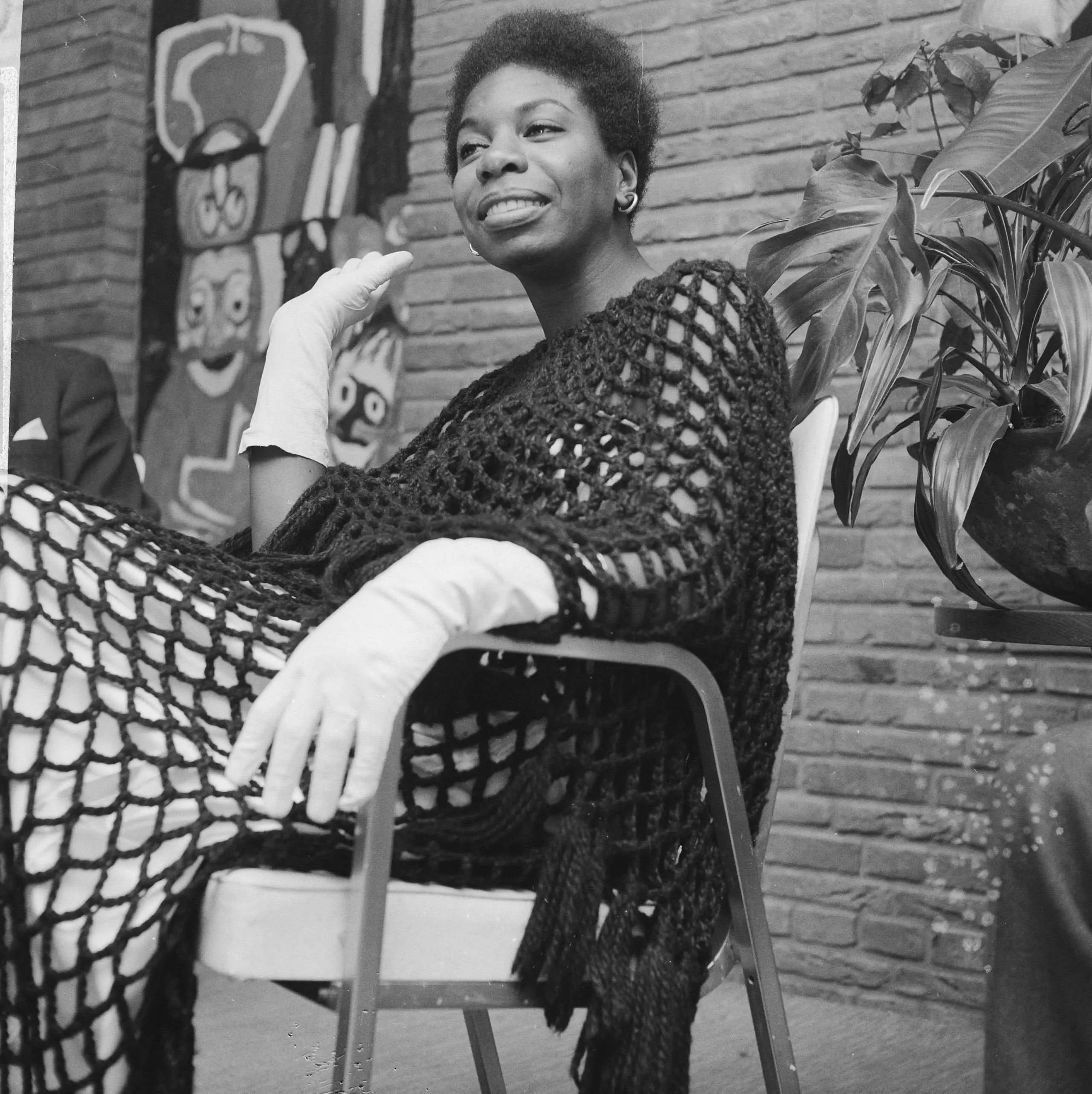 Nina Simone, wearing white gloves and an elegant crocheted tunic, lounges on a patio