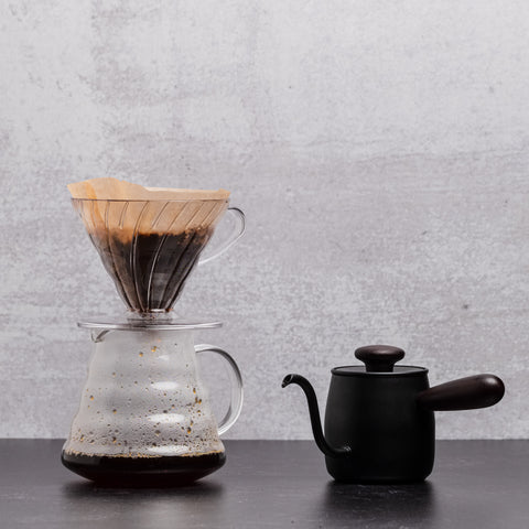 A V60 brewer sits on top of a carafe of brewed coffee with a small gooseneck kettle beside