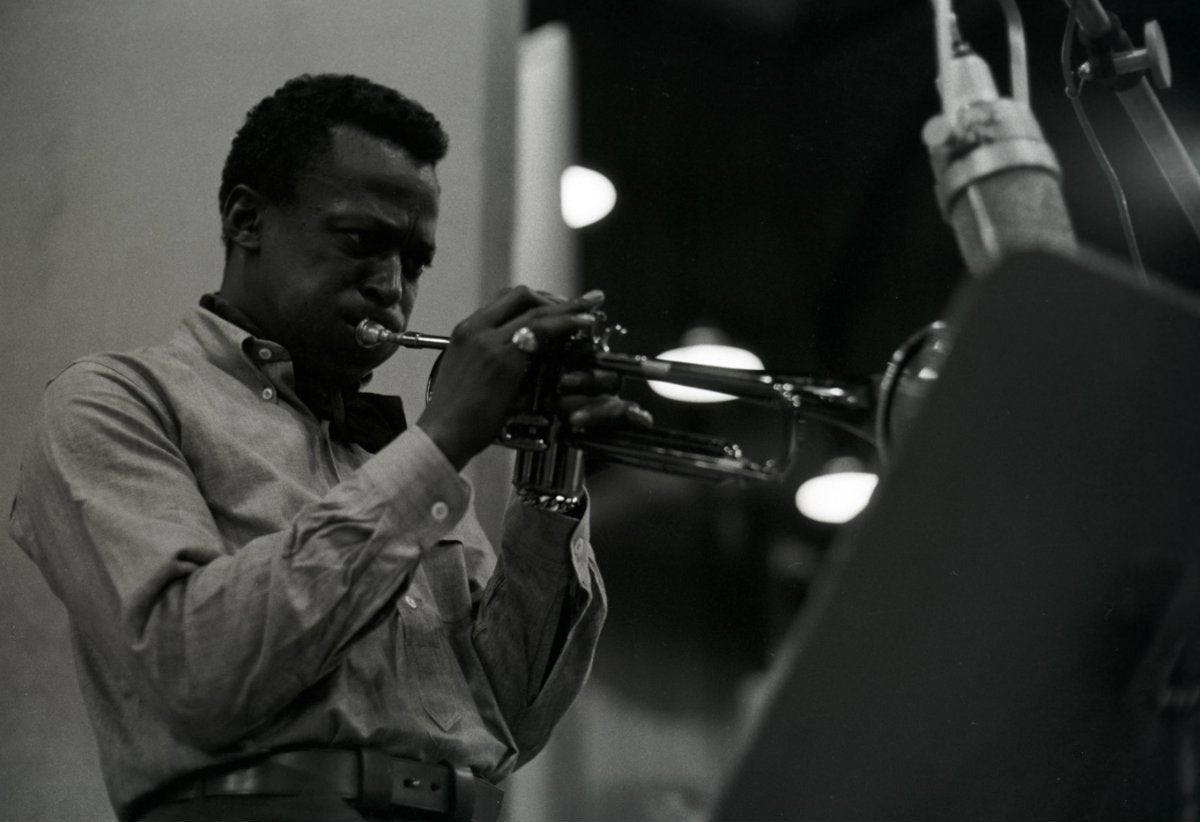 Miles Davis in a studio, playing the trumpet with a music stand and microphone in front of him