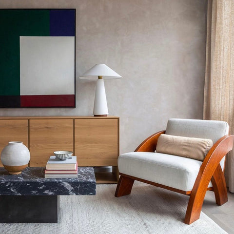 Living room with a sofa chair made of wood and white cushions, in front of  off-white curtains. Black stone coffee table in the middle of the room, sideboard plain wooden table with white lamp on top, and geometric painting on the wall.