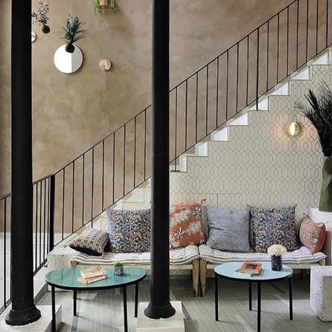 Room with bohemian-style couch against a staircase wall detailed with geometric wallpaper and 2 black pillars towards the left of the room.