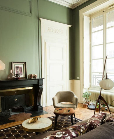 Room with pastel green walls, tall white door and white framed french window. Lounge chair and cushioned stool besides a white stone table. Black wooden fireplace.