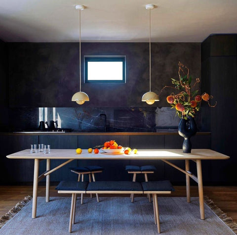 Dark purple wall with small rectangle window, wooden long table and cushioned stools.