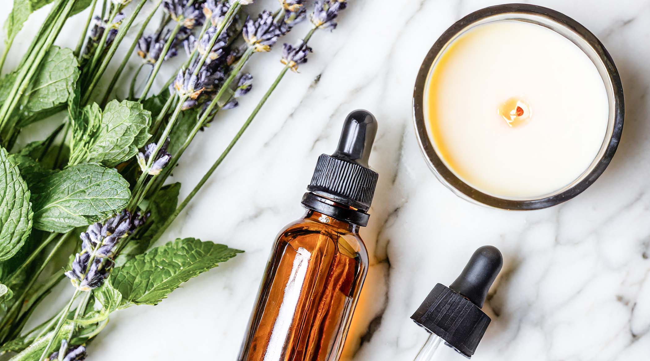 Scented soy wax blend candle with mint, lavender, and essential oil dropper bottle on marble table.