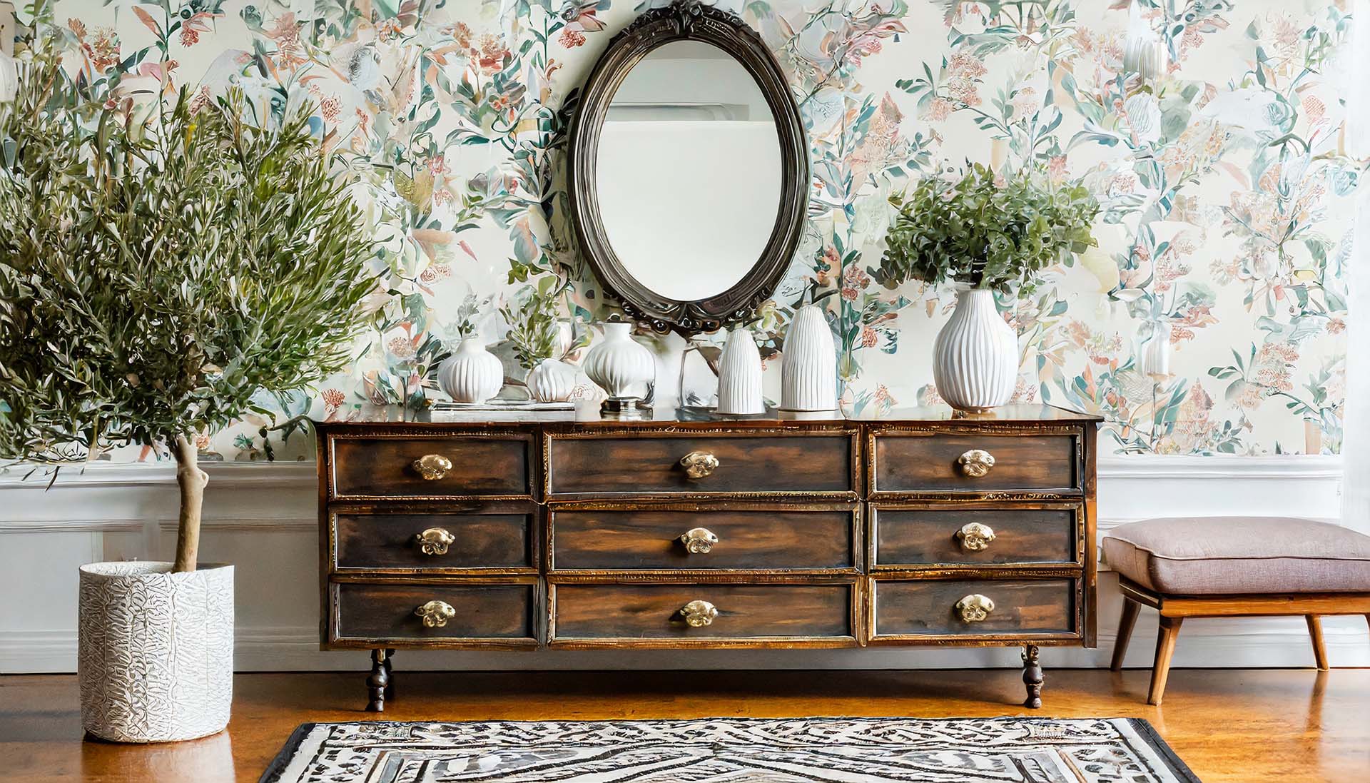 Transitional interior design with vintage apothecary chest, modern vases, traditional floral wallpaper, and olive tree.