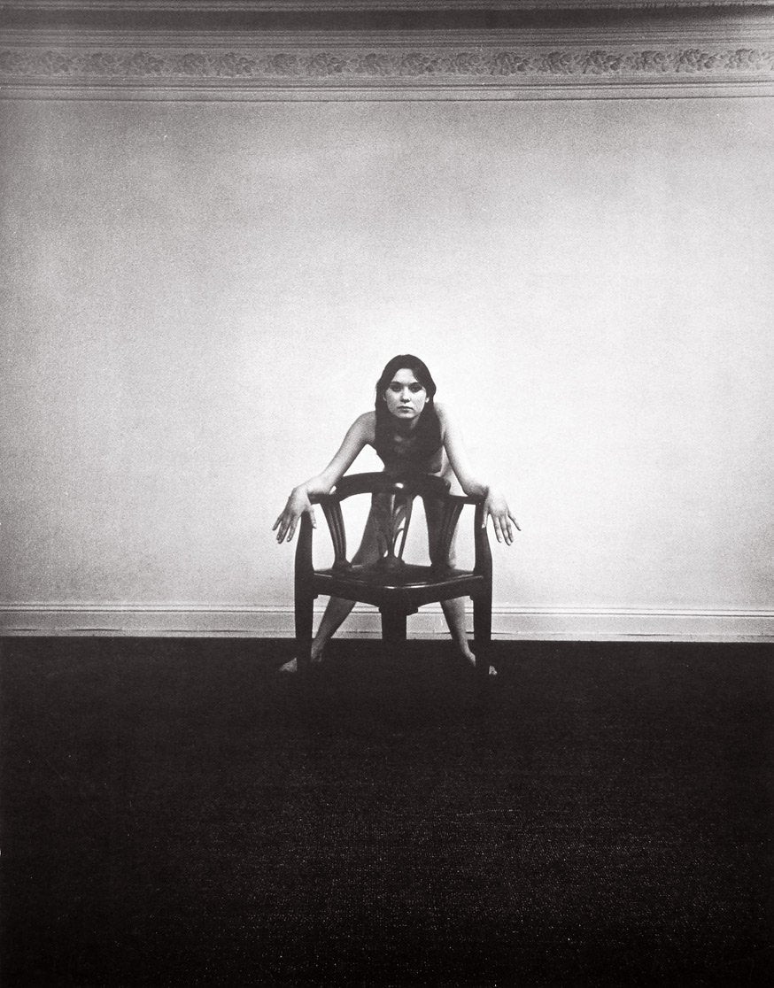 Black and white image of a nude woman leaning on a chair, facing the camera straight on