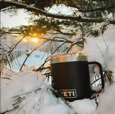 A custom YETI mug nestled in a snowbank in front of a pine tree against a backdrop of a frozen lake