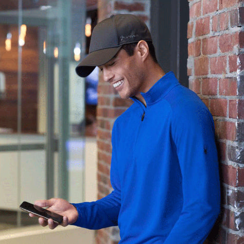 Smiling man leaning against a brick wall with a phone in hand wearing a bright blue shirt and a custom Spyder hat from Merchology
