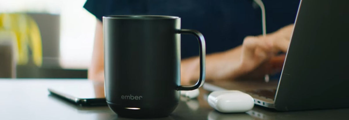A black custom logo printed Ember temperature control coffee mug sitting on a desk while a person works on a laptop