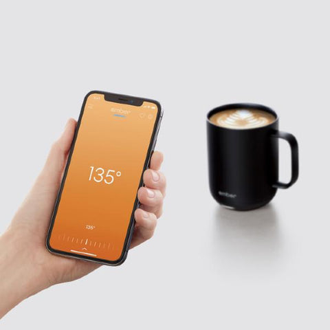 A hand holding a smartphone with a temperature reading of 135 degrees in front of a custom Ember coffee mug full of coffee